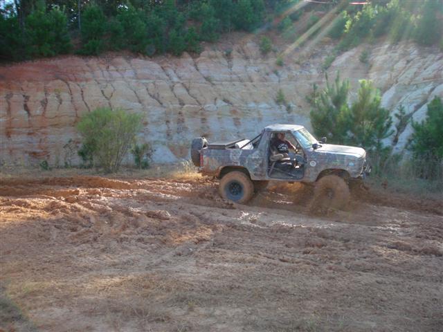 Pickup truck with camouflage paint driving through mud, driver visible. Muddy terrain and a dirt embankment in background