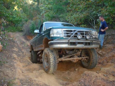 2008 International School Bus - A lifted green pickup truck navigates a muddy, off-road trail with a man standing nearby.