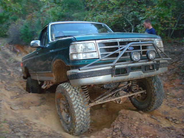 Green Ford F-Series pickup truck with a brush guard and off-road tires navigating a dirt trail