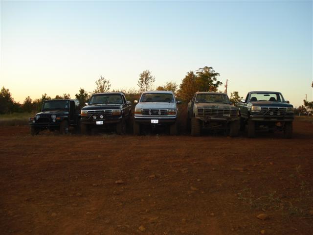 Five lifted trucks and a Jeep parked in a dirt lot under a clear sky