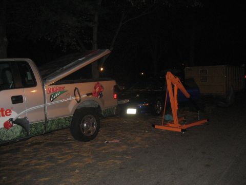 1993 Ford Probe - Pickup truck with lawn equipment graphics parked next to a car and an engine hoist at night.