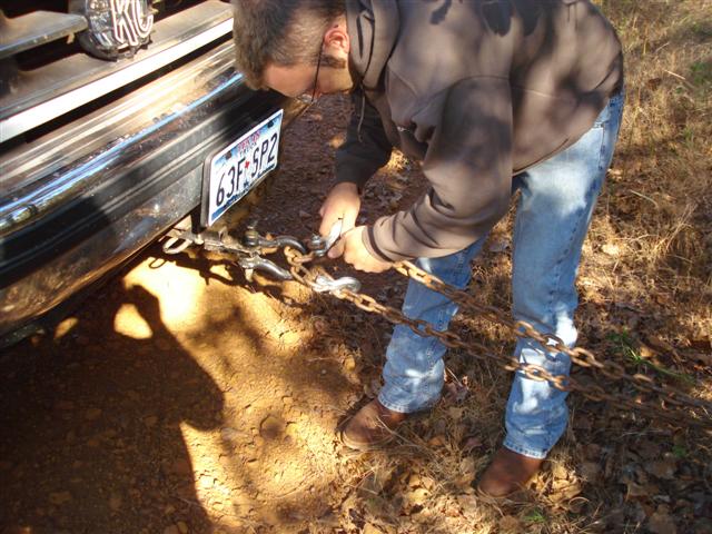 Man in jeans and hoodie attaches chains to a chrome bumper. License plate reads 63F-SP2