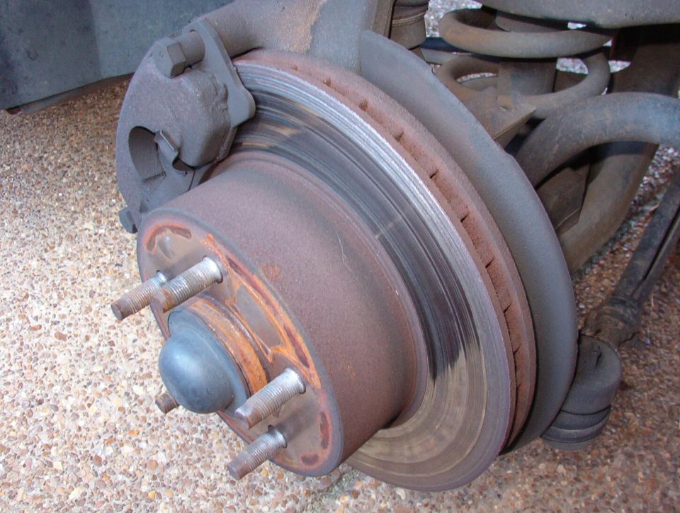 Close-up of a rusty front disc brake rotor, hub, caliper and coil spring under a vehicle