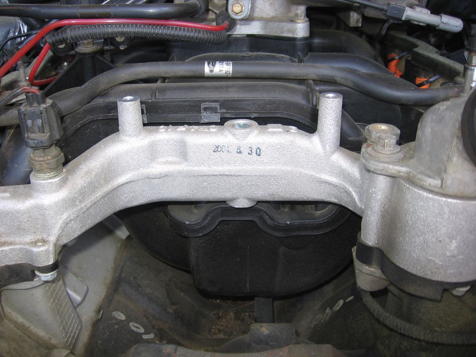 Engine bay close-up of cast aluminum bracket over black intake manifold with Ford-labeled line and wiring