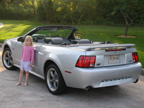 2002 Ford Mustang - Silver 2002 Ford Mustang convertible with a young girl posing next to it. The rear shows the 'MUSTANG' script and a license plate.