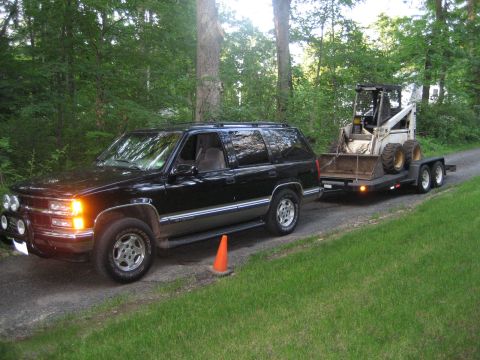 1998 Chevrolet Tahoe - A black Chevrolet Suburban from the mid-1990s is seen towing a trailer loaded with a skid steer loader on a gravel road.
