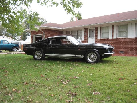 1968 Shelby GT500 - A black 1968 Shelby GT500 is parked on a lawn in front of a house, with a blue pickup truck visible in the background.