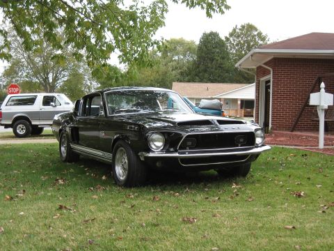 1968 Shelby GT500 - This black 1968 Shelby GT500 is captured in a three-quarter front view, parked on a grassy lawn next to a brick house.