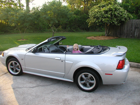2002 Ford Mustang - A silver 2002 Ford Mustang convertible is parked on a driveway with a young girl sitting in the driver's seat.