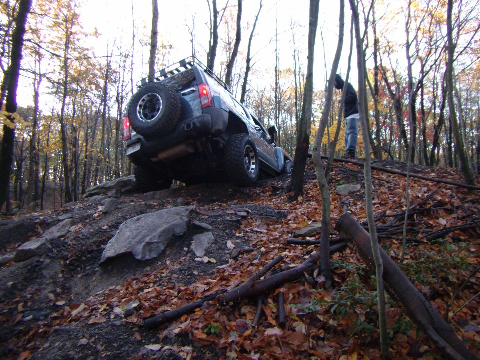 Dark blue SUV with a roof rack and spare tire on the back navigates a steep, rocky, wooded trail