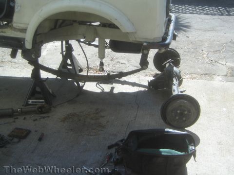 1987 Suzuki Samurai - Underside rear view of a white Suzuki Samurai up on a jack stand with its rear axle removed; the leaf spring, drum-brake backing plate, dri…