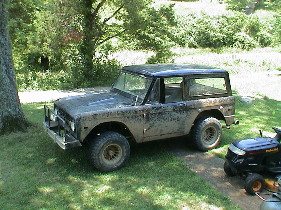 Muddy first-gen Ford Bronco parked on grass next to a riding mower