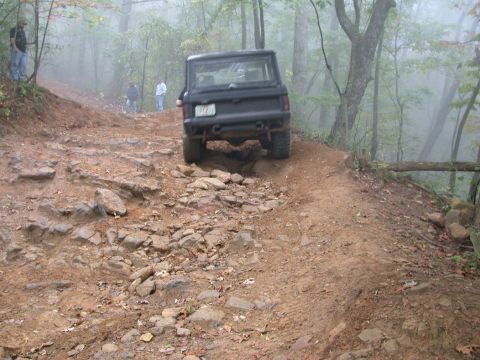 1972 Ford Bronco - A black SUV navigates a challenging, rocky off-road trail in a foggy forest setting.