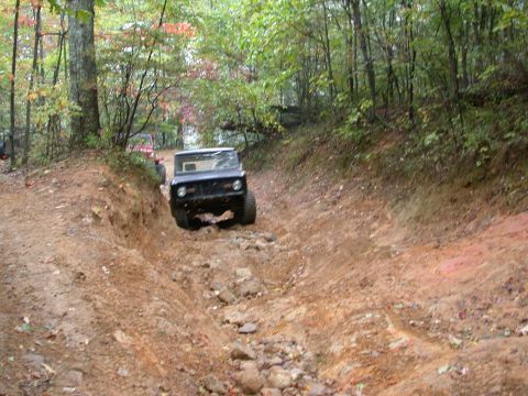 1972 Ford Bronco - A black SUV tackles a challenging off-road trail with rocky terrain and muddy inclines, surrounded by trees.