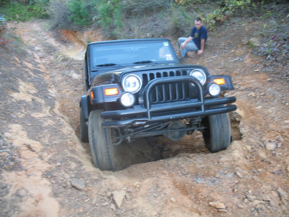 Black Jeep Wrangler with tube bumper crawling through a dirt rut, man crouching nearby