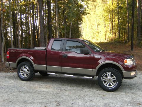 2005 Ford F-150 - Passenger-side profile of a two-tone maroon-over-tan extended-cab pickup parked on gravel in front of a sunlit pine woodland. It has tinted…