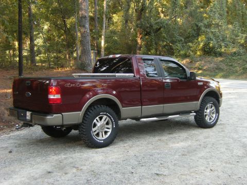 2005 Ford F-150 - A dark maroon/burgundy Ford F-150 extended-cab pickup with two-tone tan lower cladding, shown from a rear 3/4 angle on a gravel road in a w…