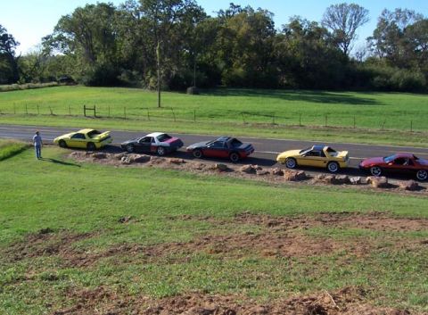 1988 Pontiac Fiero - A row of five Pontiac Fieros, including yellow, red, and two-tone models, are parked along a grassy roadside.