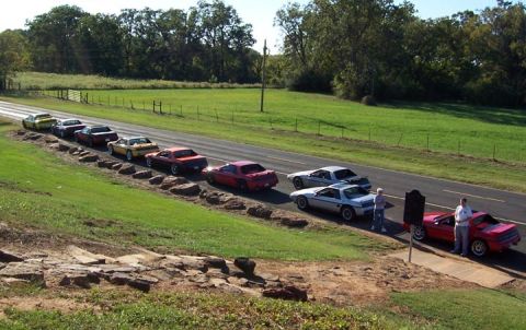 1988 Pontiac Fiero - A group of 1988 Pontiac Fieros are lined up on the side of a road with people standing near them.