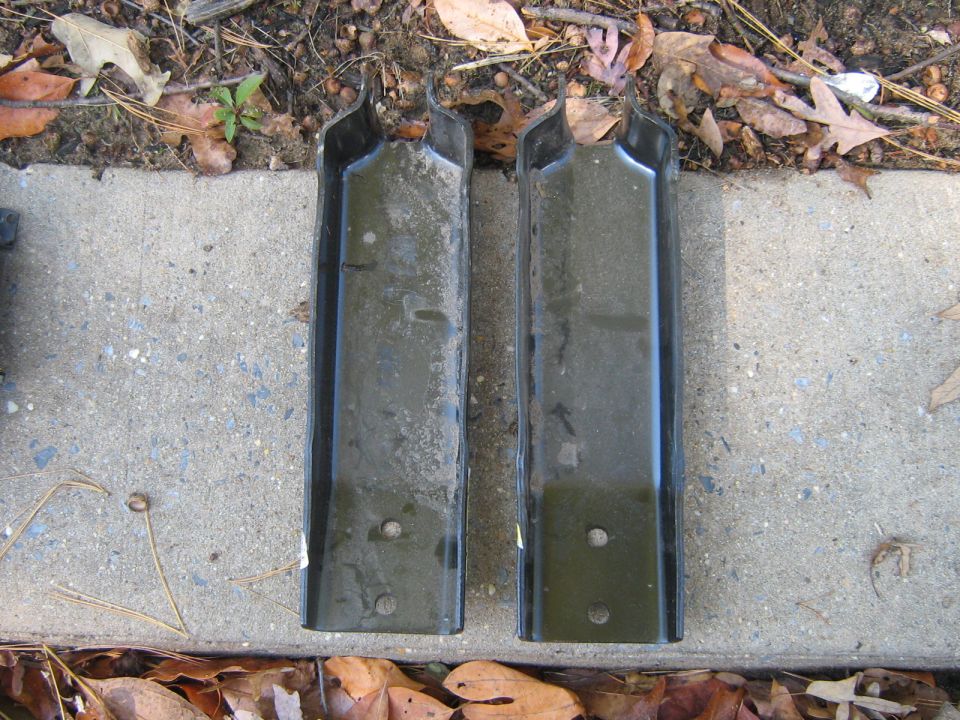 two black metal brackets with holes, lying on concrete and dirt