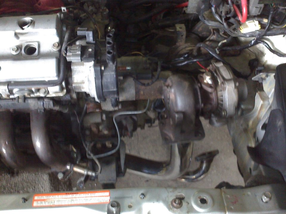 Engine bay showing turbocharger, exhaust manifold, and wiring. Orange sticker with text is visible