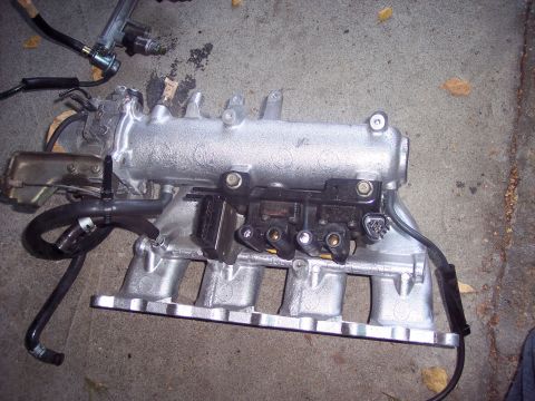 1995 Mitsubishi Eclipse - A silver intake manifold with attached hoses and electrical components is shown on a concrete surface.