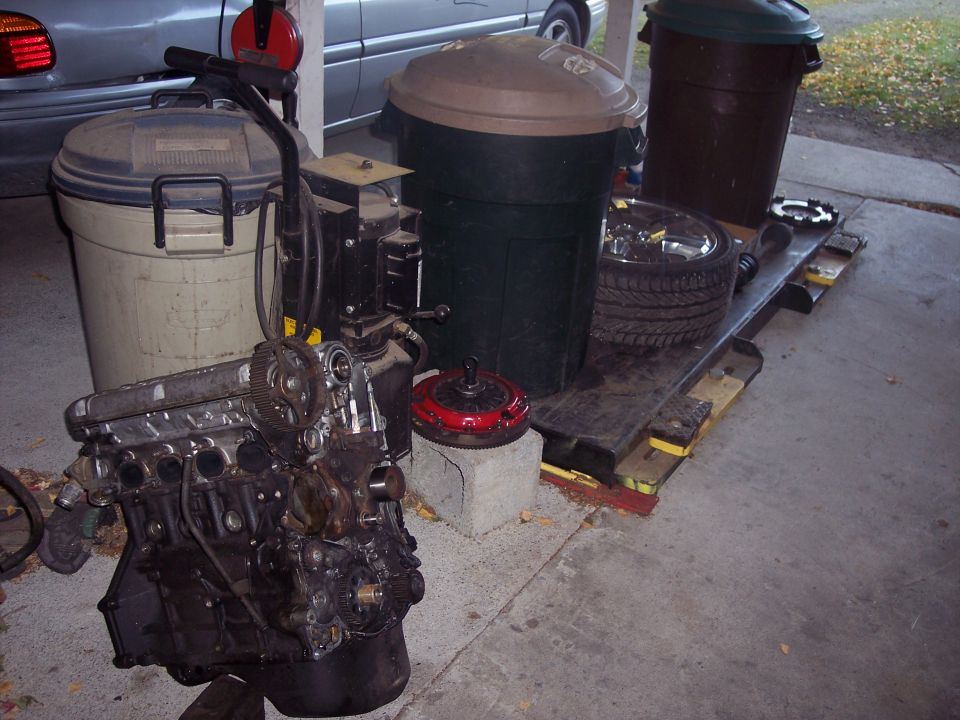 Engine block and red torque converter on concrete floor with trash cans and a tire in background