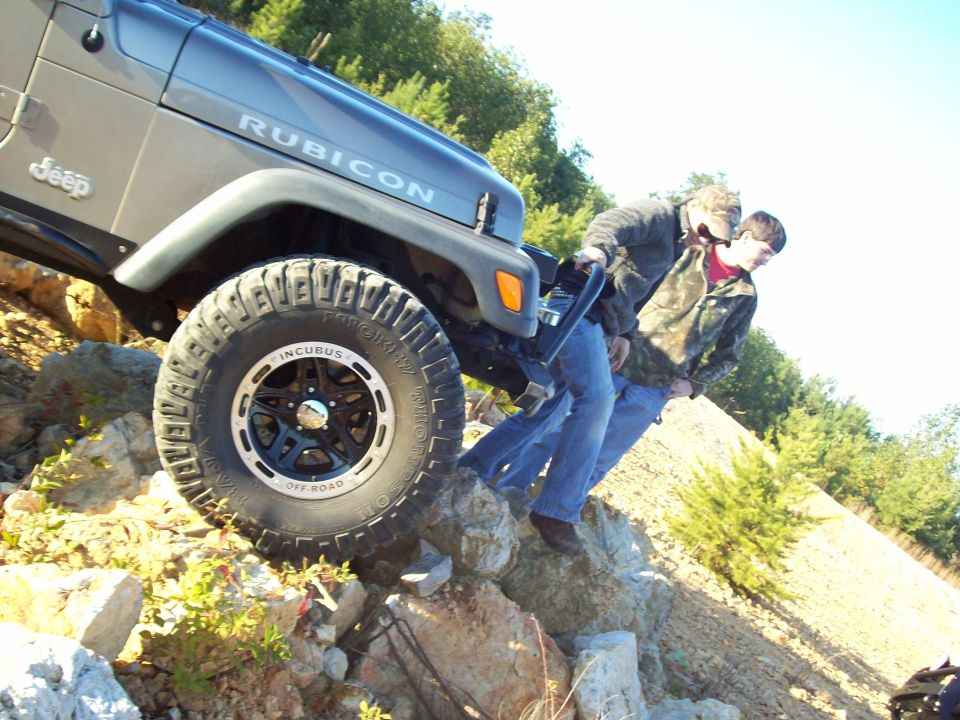 Jeep Wrangler Rubicon with 'INCUBUS OFF-ROAD' wheels on rocky terrain, two people pushing