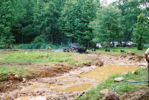 1972 Ford Bronco - A dark SUV navigates a muddy track with trees in the background and other vehicles parked further away.