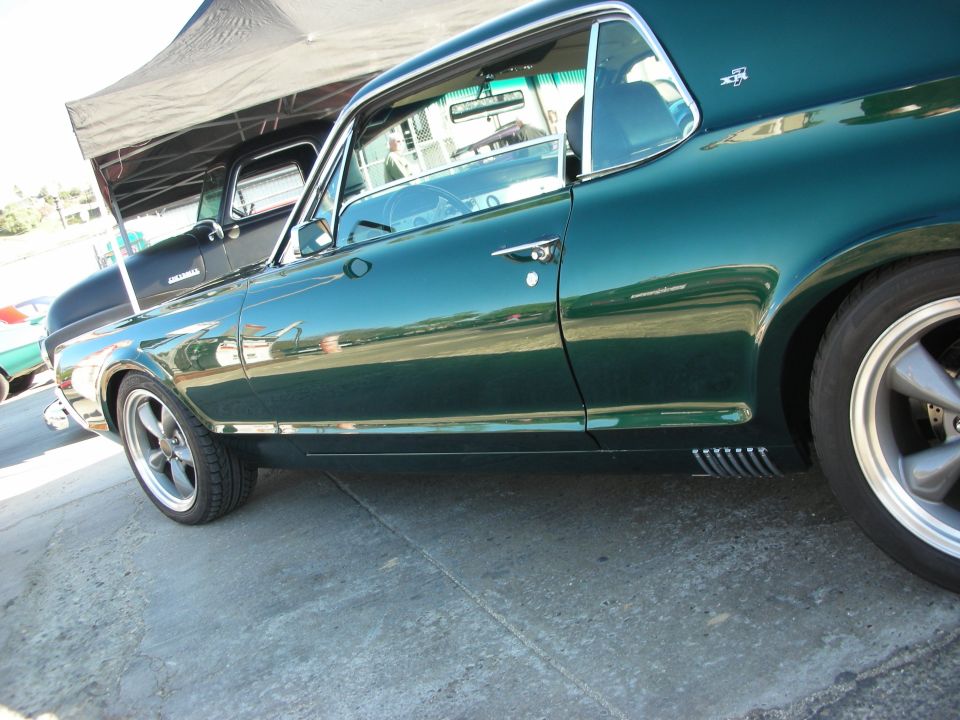 Side view of a dark green 1967 Mercury Cougar coupe with aftermarket wheels. A black pickup truck is parked behind it