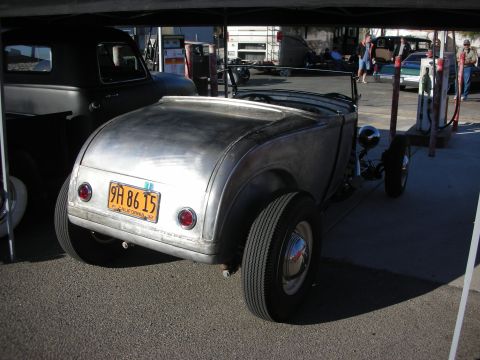 1998 Ford Crown Victoria - A bare metal hot rod is parked next to a black pickup truck at a gas station.