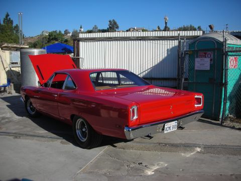 1998 Ford Crown Victoria - A red 1969 Plymouth Road Runner coupe is parked with its hood up next to a portable toilet.