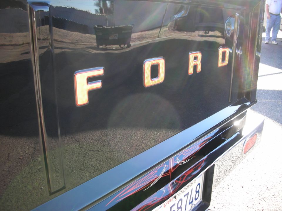 Black Ford truck tailgate with chrome and orange lettering spelling "FORD". Pinstriping on bumper