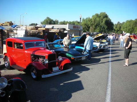 1998 Ford Crown Victoria - A red sedan hot rod is parked at a car show, with other vehicles and spectators in the background.