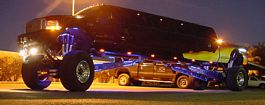 1992 Ford Bronco - A custom-built monster truck limo with a yellow pickup truck loaded on top is illuminated by blue lights at night.