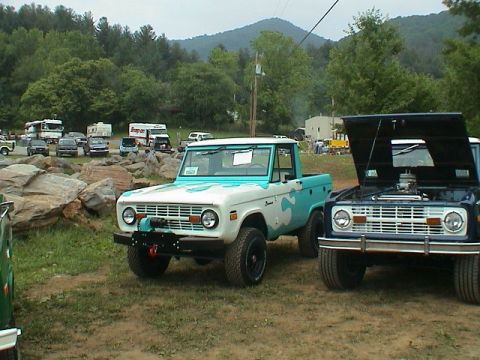 1972 Ford Bronco - Two Ford Broncos are parked at an outdoor event. The one in front has turquoise camouflage paint and a winch.