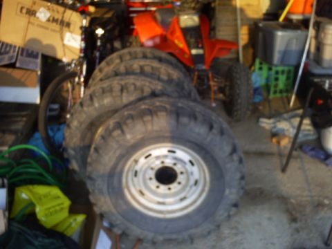 Four large, knobby tires with silver rims stacked in a garage. A red ATV is visible in the background