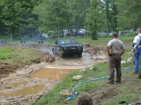 1972 Ford Bronco - A dark-colored off-road vehicle navigates a challenging muddy course with spectators watching from the side.