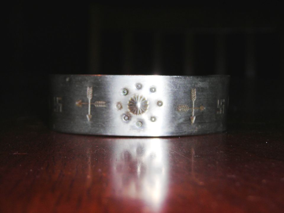 Silver metal bracelet with etched arrows and a central sunburst design, resting on a wooden surface