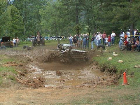 1972 Ford Bronco - A crowd watches as a modified off-road vehicle navigates a deep mud pit, kicking up a large spray.