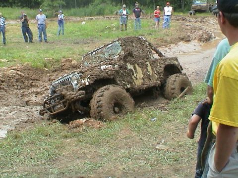 1972 Ford Bronco - A Jeep is stuck deep in mud, with onlookers observing the scene.