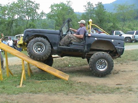 1972 Ford Bronco - A black convertible SUV is positioned on a yellow ramp with a driver inside, while other vehicles are parked in the background.