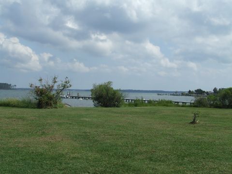 1976 Ford F-150 - A wide shot of a calm body of water with a wooden pier extending into it, bordered by green grass and trees.