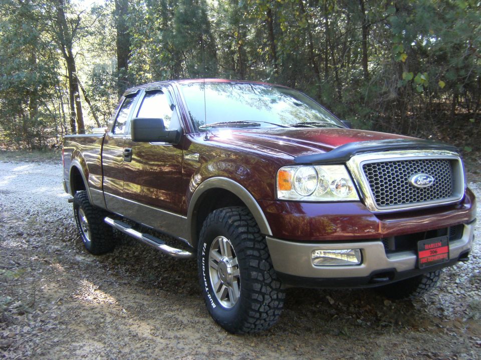 Maroon Ford F-150 extended-cab pickup, front 3/4 view, on gravel in woods with mud tires