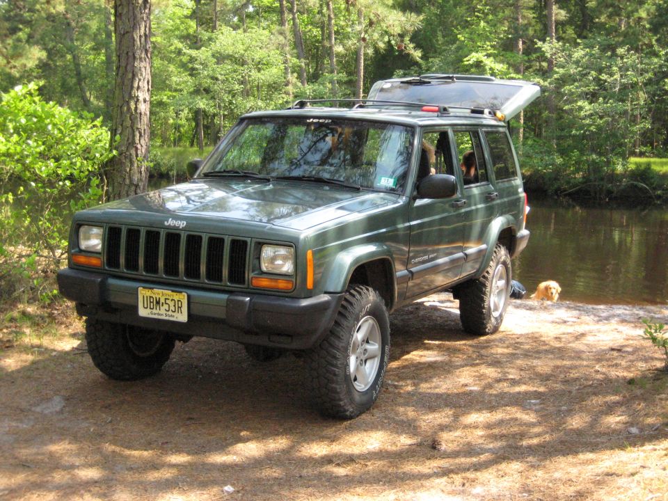 Green lifted Jeep Cherokee XJ, front 3/4 view, parked by water in woods