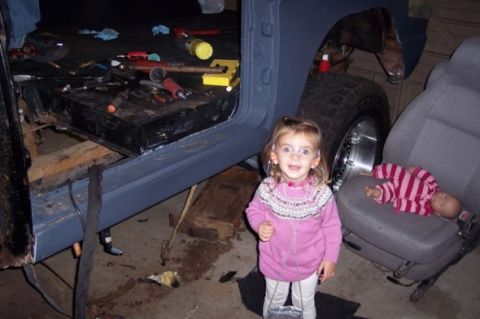 1976 Ford Bronco - A young child stands in a garage next to a vehicle undergoing repairs, with tools and parts visible.