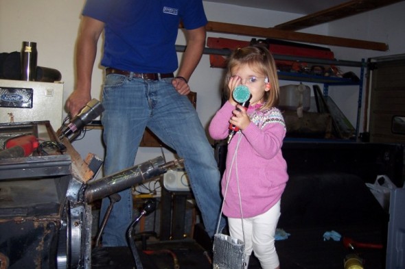 Girl holding a microphone in a garage with a man and car parts