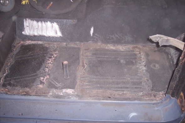Vehicle floor with rust and white paint marks, showing a metal post and torn fabric