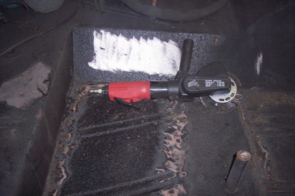 Air grinder with wire wheel attachment on a dark, textured surface, likely a vehicle floor undergoing rust removal