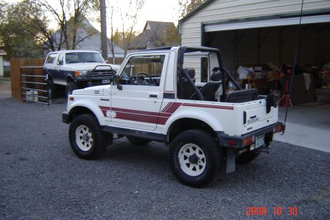 1988 Ford Bronco - A white Suzuki Samurai with red side stripes is parked in a gravel driveway next to a garage, with another SUV in the background.